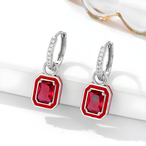A pair of silver hoop earrings with a line of small diamonds on the front, featuring square-cut red gemstones in a silver setting, resting on a white surface.
