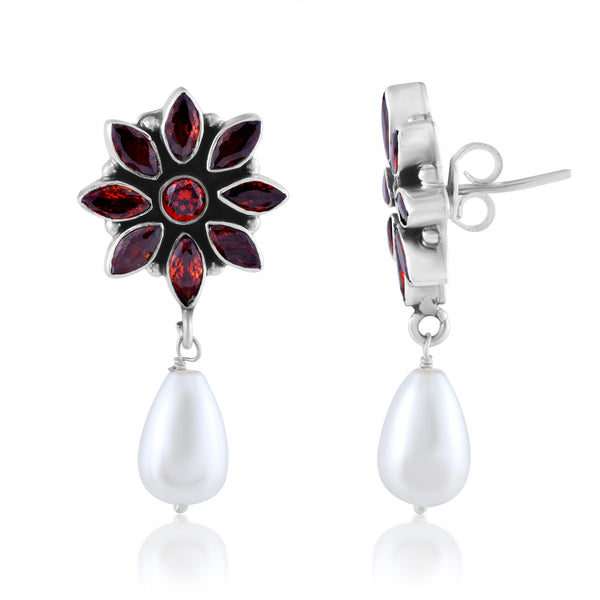 A pair of silver dangle earrings with a flower-shaped top adorned with red gemstones, and a dangling white teardrop-shaped pearl.