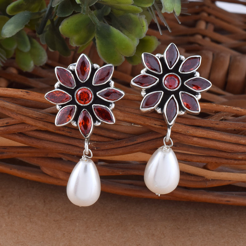 A pair of silver earrings with flower-shaped studs encrusted with red gemstones and dangling white teardrop pearls, displayed on a woven twig surface with some green leaves in the background.