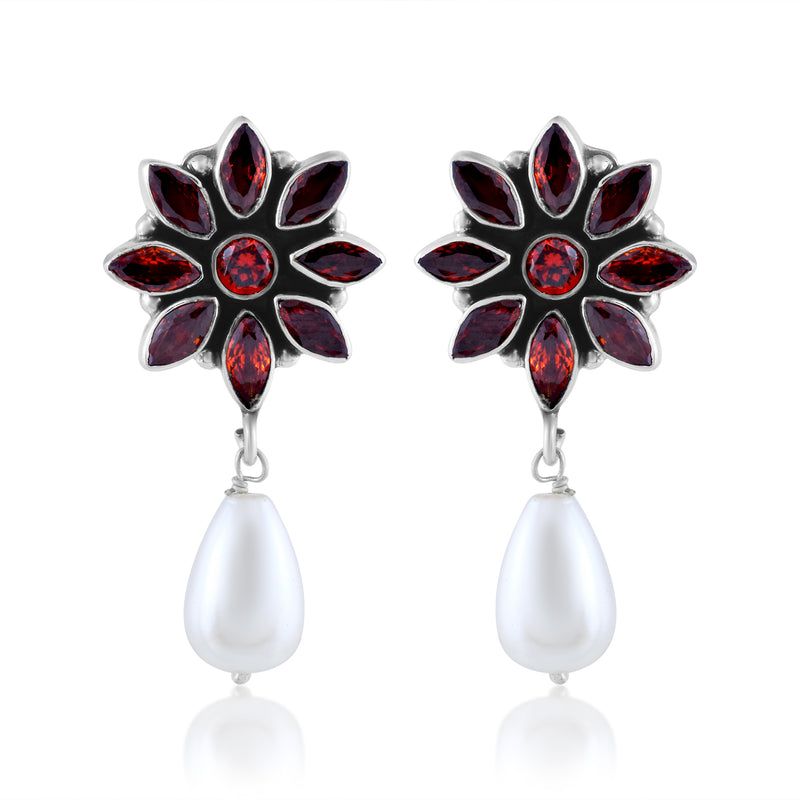 A pair of silver earrings with a flower design made of red gemstones and a dangling white pearl.