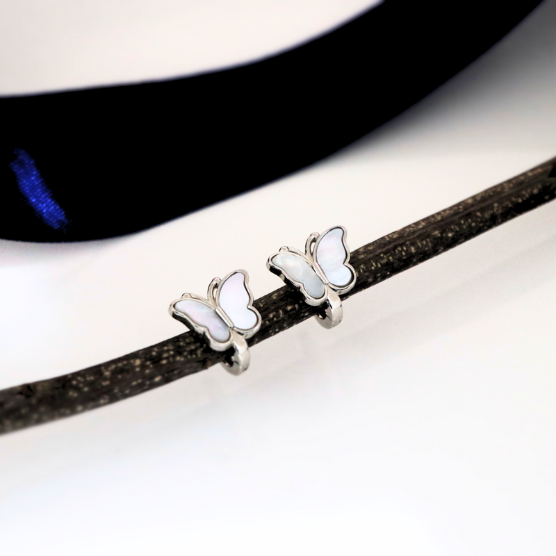 A pair of silver butterfly earrings with white wings resting on a dark textured surface with a blurred circular object in the background.